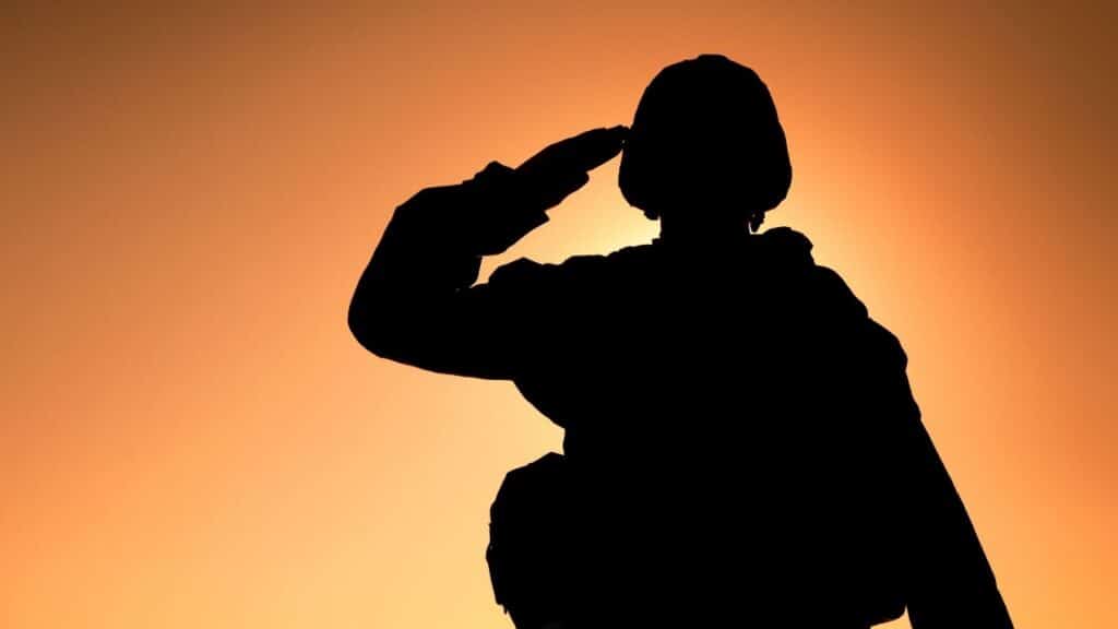 A dramatic, silhouetted image of a soldier or service member in uniform and helmet rendering a hand salute against a bright orange and yellow sunset or sunrise sky.