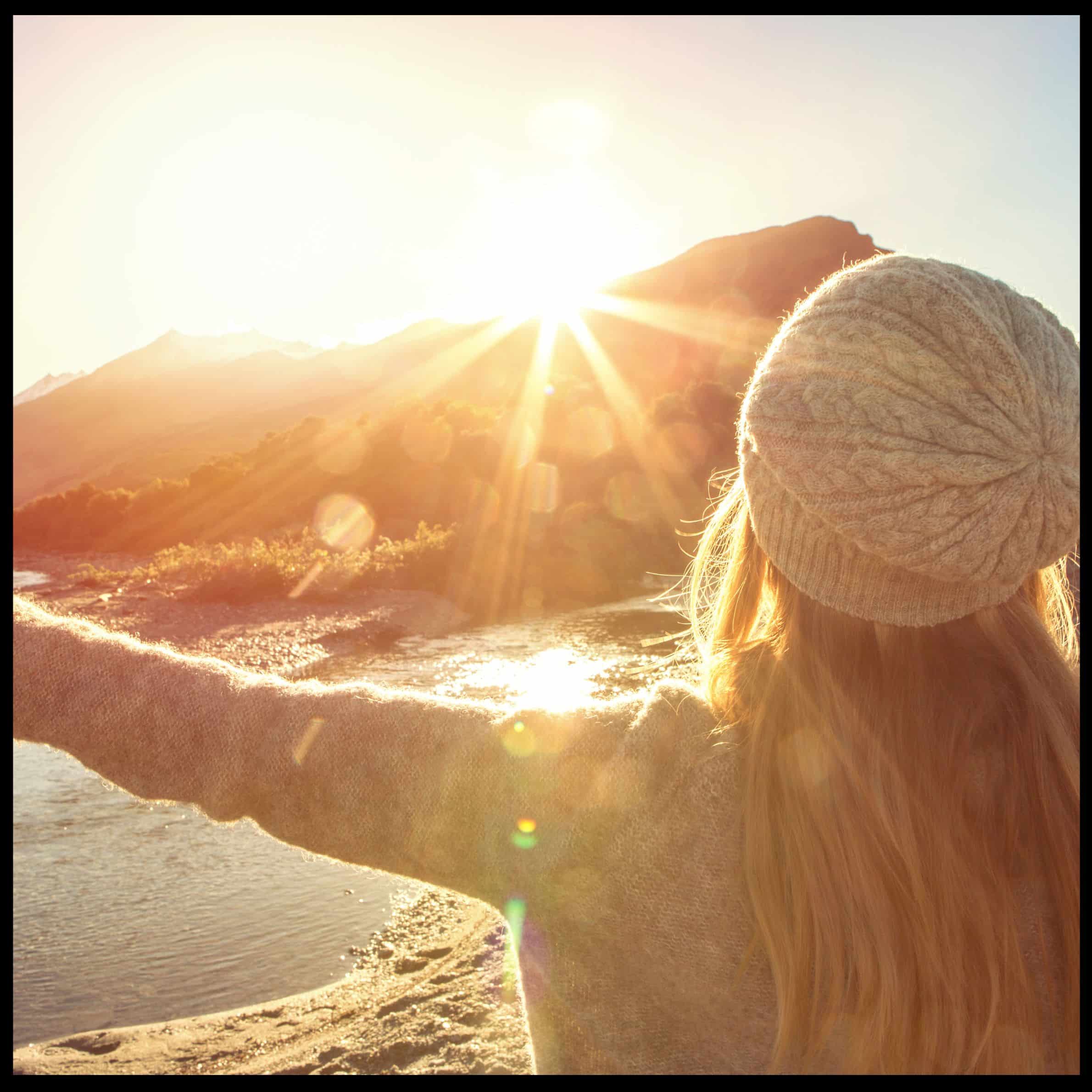 A woman in a beanie faces a brilliant sunrise over mountains and a river, with an arm outstretched.